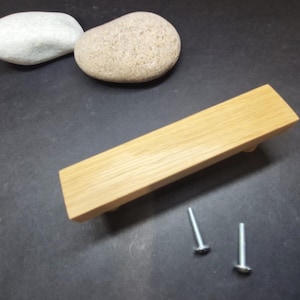 May include: A rectangular light brown wooden cabinet handle with a visible grain pattern, featuring a flat top surface and angled sides. Two silver-coloured metal screws are positioned separately near the handle for installation purposes.