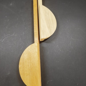 May include: Two light-coloured wooden kitchen utensils, each featuring a long, straight handle attached to a semi-circular head. The wood grain is visible on the flat surfaces of the light tan, natural-toned wooden spoons or spatulas.