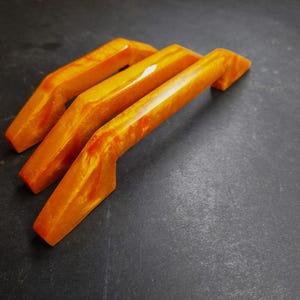 May include: Three cabinet handles with a glossy, semi-translucent finish in shades of vibrant orange and yellow. Each handle features an angular, geometric design with a smooth, polished surface that reflects light, showcasing a marbled color pattern.