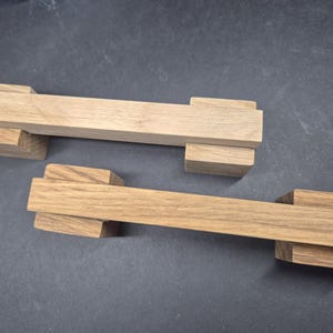 May include: Two light brown wooden handles, each featuring a long, rectangular central bar supported by two smaller, block-shaped wooden pieces at either end. The wood grain is visible on the surface of both handles, which are positioned diagonally.