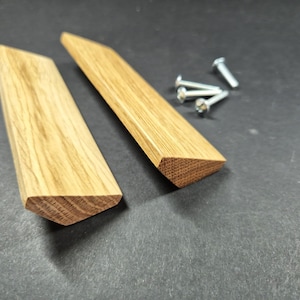 May include: Two light brown wooden cabinet pulls with a triangular profile and visible wood grain, accompanied by four small silver metal screws. The pulls have a smooth finish and a distinct angled shape, designed for attachment to furniture doors.