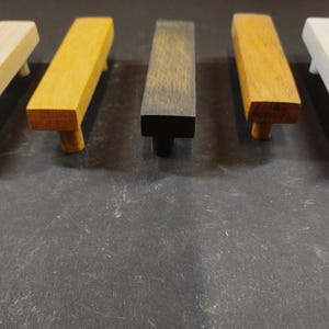 Puede incluir: Cinco tiradores rectangulares de madera para armarios dispuestos en fila. Los tiradores presentan diferentes acabados, incluyendo madera natural clara, roble dorado, carbón oscuro, marrón medio y un blanco roto pálido, cada uno con dos postes de montaje.