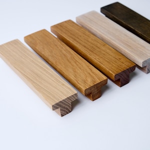 Op de afbeelding: Vijf houten kastgrepen in verschillende bruintinten, van licht tot donker. De grepen hebben allemaal dezelfde vorm, met een rechthoekig lichaam en een T-vormig uiteinde.