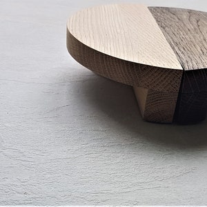 Può includere: Un piedistallo rotondo in legno con una finitura in legno chiaro e scuro. Il piedistallo è composto da due metà, una chiara e una scura, che sono unite. Il piedistallo ha un diametro di circa 25 cm.