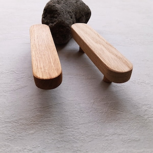 Könnte beinhalten: Zwei Holzknaufgriffe mit abgerundeter rechteckiger Form. Die Griffe sind aus hellem braunem Holz gefertigt und haben eine glatte Oberfläche.