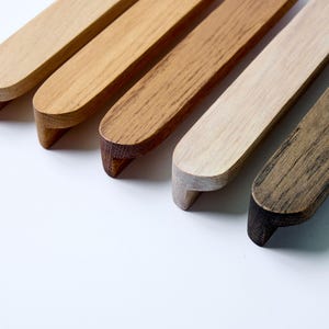 May include: Five wooden drawer pulls in different wood finishes, including natural, light brown, dark brown, and grey. The pulls are all the same shape, with a rounded edge and a flat bottom.