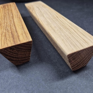 May include: Two elongated wooden blocks with a trapezoidal cross-section. The left block features a medium-brown wood grain, while the right block is a lighter, pale beige wood. Both pieces display a visible natural wood texture and grain pattern.