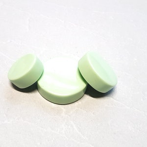 May include: Three pale green, circular soap bars are displayed on a light grey surface. Two bars are angled, and one is flat. The soap has a smooth, solid appearance, suggesting a handmade or natural product. The image is well-lit, highlighting the colour and shape.