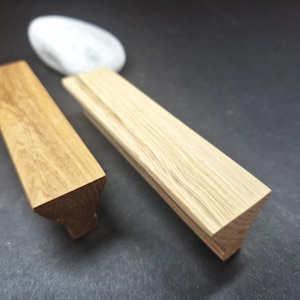 May include: Two elongated wooden cabinet pulls with a trapezoidal profile. One pull features a medium brown wood grain, while the other is a lighter, pale wood tone. Both handles have a flat top surface and angled sides that taper inward.