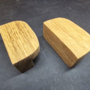 May include: Two semi-circular wooden cabinet pulls with a natural light brown wood grain finish. Each handle features a flat front edge and a curved top profile, showcasing the organic texture and vertical grain pattern of the light oak wood.