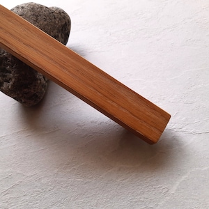 May include: A long, rectangular wooden drawer pull with a natural wood finish. The pull is lying on a white textured surface.