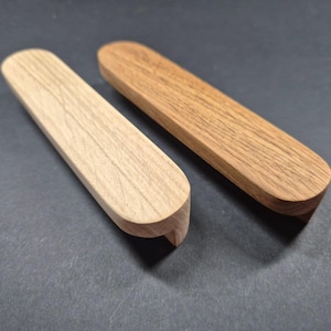 Puede incluir: Dos tiradores de cajón rectangulares de madera con extremos redondeados. Uno está hecho de madera clara con veta visible, mientras que el otro es de madera más oscura con tonos cálidos. Ambos presentan un acabado liso y un diseño curvo.