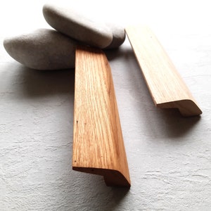 May include: Three wooden cabinet door pulls with a rounded rectangular shape and a slight curve at the bottom. The pulls are made of light brown wood with a natural grain pattern.