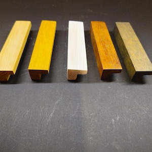 Puede incluir: Cinco tiradores de armario rectangulares de madera dispuestos en fila. Los tiradores presentan acabados distintos que incluyen madera natural clara, amarillo brillante, blanco, marrón medio y verde oliva oscuro, con perfil en L y veta.