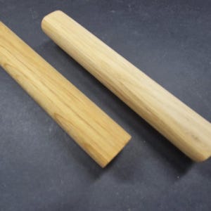 May include: Two cylindrical wooden dowels with a smooth, light tan finish and visible natural wood grain patterns. Each piece features rounded ends and a uniform, straight shape, showcasing the raw texture and warm color of the light-toned wood.
