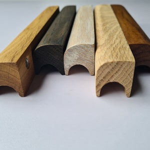 Könnte beinhalten: Fünf Holzknaufgriffe in verschiedenen Holztönen, darunter natur, weiß getönt, dunkel gebeizt und rotbraun. Die Griffe haben alle die gleiche Form, mit einer abgerundeten Oberseite und einer geschwungenen Unterseite.