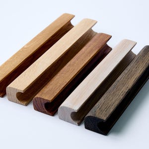 Puede incluir: Cinco tiradores de cajón de madera en diferentes tonos de marrón, desde claro a oscuro. Los tiradores tienen todos la misma forma, con una base rectangular y una parte superior curva.