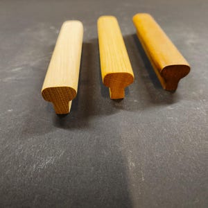 Könnte beinhalten: Drei Schrankgriffe aus Holz, die in einer Reihe angeordnet sind und jeweils ein T-förmiges Profil mit einer abgerundeten Oberkante aufweisen. Die Griffe zeigen unterschiedliche Schattierungen der natürlichen Holzmaserung, von einem hellen Ton bis zu einem wärmeren Braun.