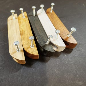 Könnte beinhalten: Fünf längliche Schubladengriffe aus Holz, die in einer Reihe angeordnet sind. Jeder Griff hat ein geschwungenes Profil und zwei silberfarbene Metallschrauben, die oben herausragen. Die Holzmaserung variiert von hellem Eichenholz bis zu Anthrazit.