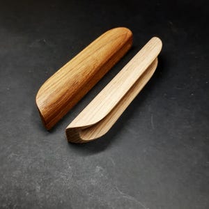 May include: Two wooden drawer pulls, one with a rounded edge and the other with a curved edge. Both pulls are made of light brown wood.