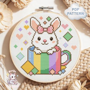 May include: A cross stitch pattern in a wooden embroidery hoop featuring a white bunny with a pink polka dot bow sitting inside a rainbow striped mug. The design includes small colorful hearts and diamond shapes on a white fabric background.