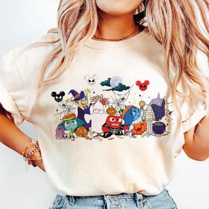 Peut inclure: T-shirt crème avec un motif d'Halloween. Le design comprend des personnages de dessins animés costumés, des araignées, des toiles d'araignées et des silhouettes de Mickey Mouse. Les personnages sont de différentes couleurs, dont le bleu, le violet, l'orange et le vert.