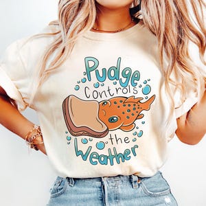 May include: A white t-shirt featuring a cartoon illustration of a fish wearing a peanut butter and jelly sandwich. The text on the shirt reads "Pudge Controls the Weather".