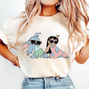 May include: A white t-shirt with a cartoon image of Stitch and Lilo from the Disney movie Lilo & Stitch. Stitch is wearing sunglasses and a green lei, while Lilo is wearing a pink dress and sunglasses. The shirt is a light ivory color.