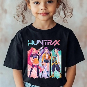 May include: Black t-shirt featuring the word "HUNTRAX" in a colorful font above a graphic of three anime-style characters. The characters are in various poses and outfits. The t-shirt is worn by a young girl.
