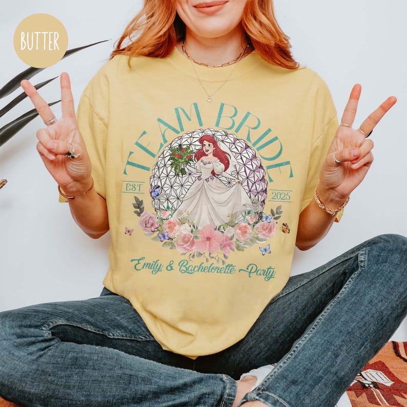 May include: Yellow t-shirt with a green and purple graphic of a Disney princess in front of a sphere. The text "Team Bride Est 2025" is on the shirt. The text "Emily & Bachelorette Party" is also on the shirt.