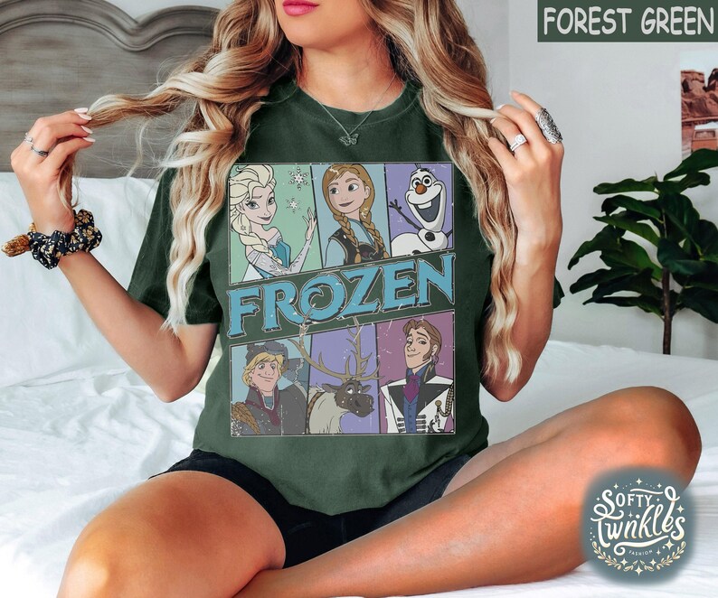 May include: A forest green t-shirt with a graphic print of characters from the movie Frozen. The graphic includes Elsa, Anna, Kristoff, Olaf, and Sven. The text "FROZEN" is printed in white letters on the shirt.