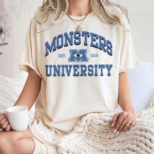 May include: Cream-colored t-shirt featuring the text "MONSTERS UNIVERSITY" in blue, with a stylized eye logo. The shirt has short sleeves and a relaxed fit, perfect for casual wear.