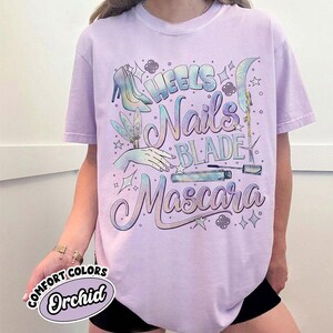 May include: A light purple t-shirt with the words "Heels, Nails, Blade, Mascara" in a decorative font. The shirt features illustrations of high heels, a hand, a nail file, and mascara. The shirt also has the words "Comfort Colors" and "Orchid" on it.