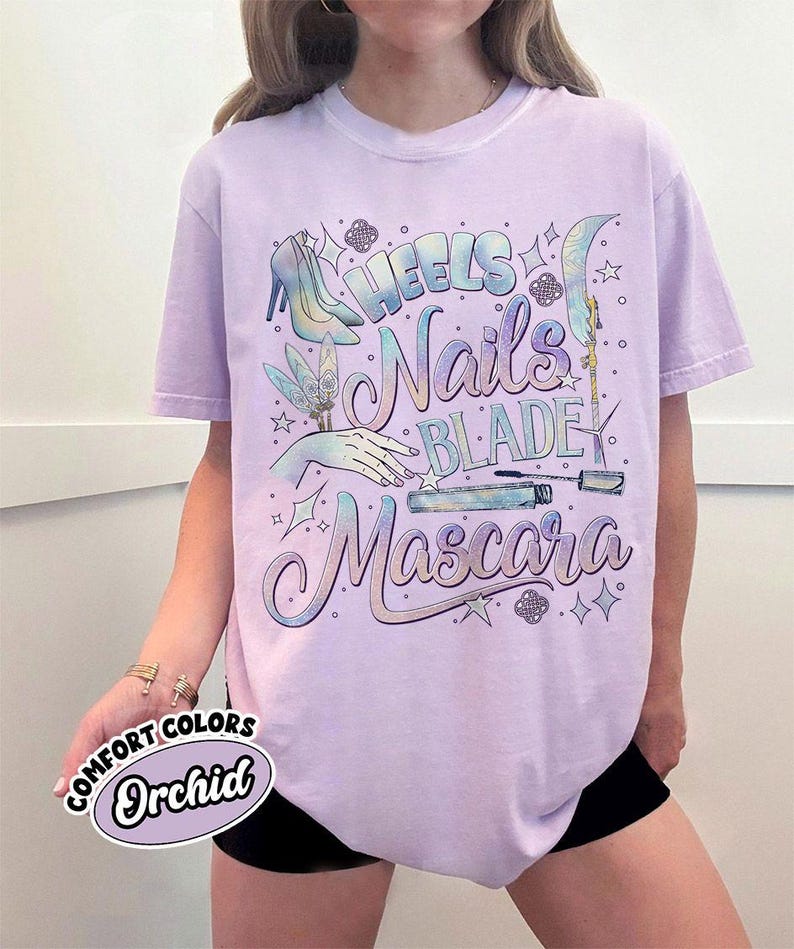 May include: A light purple t-shirt with the words "Heels, Nails, Blade, Mascara" in a decorative font. The shirt features illustrations of high heels, a hand, a nail file, and mascara. The shirt also has the words "Comfort Colors" and "Orchid" on it.