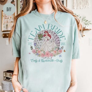 May include: A light blue t-shirt with the text "Team Bride Est 2025" and a graphic of Ariel from The Little Mermaid in a wedding dress inside a floral wreath. The text "Emily & Bachelorette Party" is below the graphic.