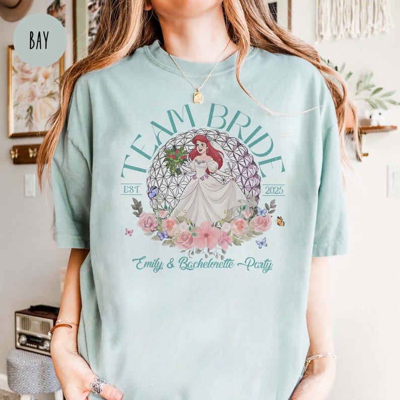 May include: A light blue t-shirt with the text "Team Bride Est 2025" and a graphic of Ariel from The Little Mermaid in a wedding dress inside a floral wreath. The text "Emily & Bachelorette Party" is below the graphic.