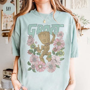 May include: A light blue t-shirt featuring a floral design and a graphic of Groot from Guardians of the Galaxy. The text "Groot" is printed in large letters above the graphic.