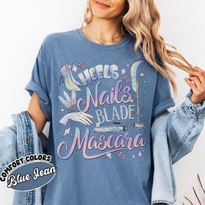 May include: Blue jean colored t-shirt with the words "Heels, Nails, Blade, Mascara" in a holographic design. The shirt also features illustrations of high heels, a hand, a blade, and mascara. The words "Comfort Colors" and "Blue Jean" are printed on a circular label.