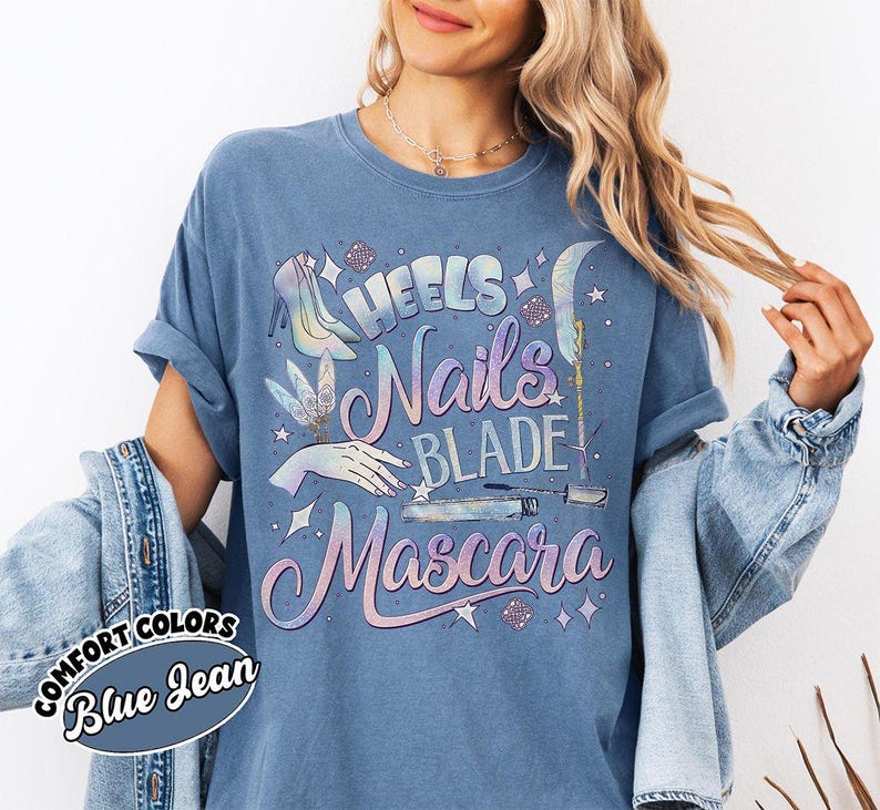 May include: Blue jean colored t-shirt with the words "Heels, Nails, Blade, Mascara" in a holographic design. The shirt also features illustrations of high heels, a hand, a blade, and mascara. The words "Comfort Colors" and "Blue Jean" are printed on a circular label.