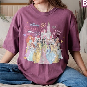 May include: A purple t-shirt with a graphic of Disney princesses in front of Cinderella's castle. The text "Disney Princess" is printed on the shirt.