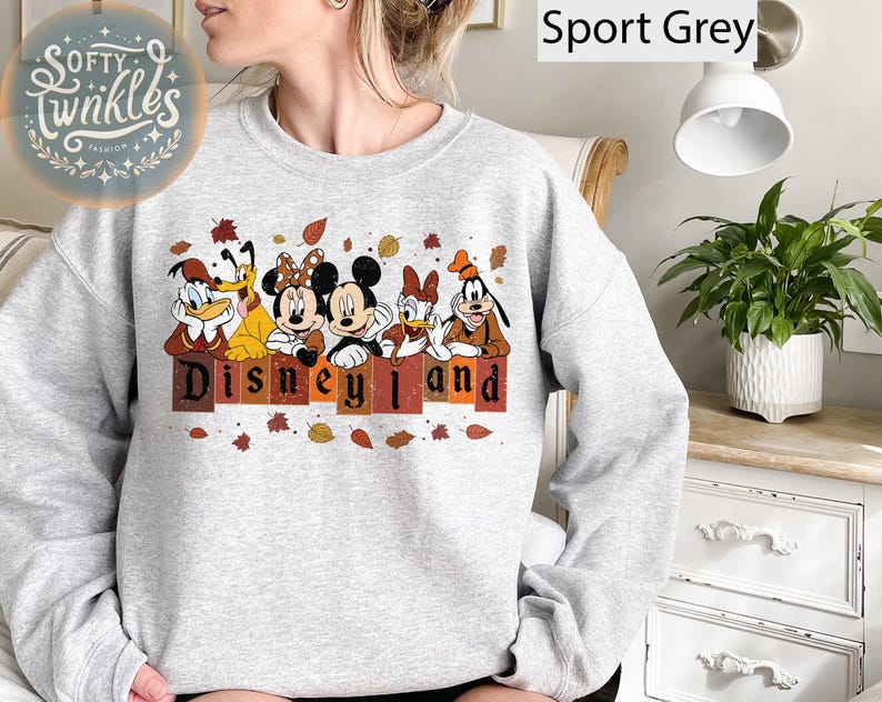 May include: A gray sweatshirt with a print of Mickey Mouse, Donald Duck, Goofy, and Pluto in front of a sign that says "Disneyland". The characters are wearing fall-themed outfits and there are leaves scattered around them.