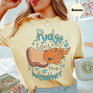 May include: A yellow t-shirt featuring a cartoon illustration of a fish with a sandwich on its head. The text "Pudge controls the weather" is printed on the shirt.