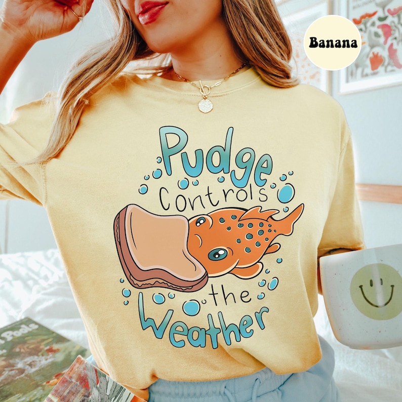 May include: A yellow t-shirt featuring a cartoon illustration of a fish with a sandwich on its head. The text "Pudge controls the weather" is printed on the shirt.