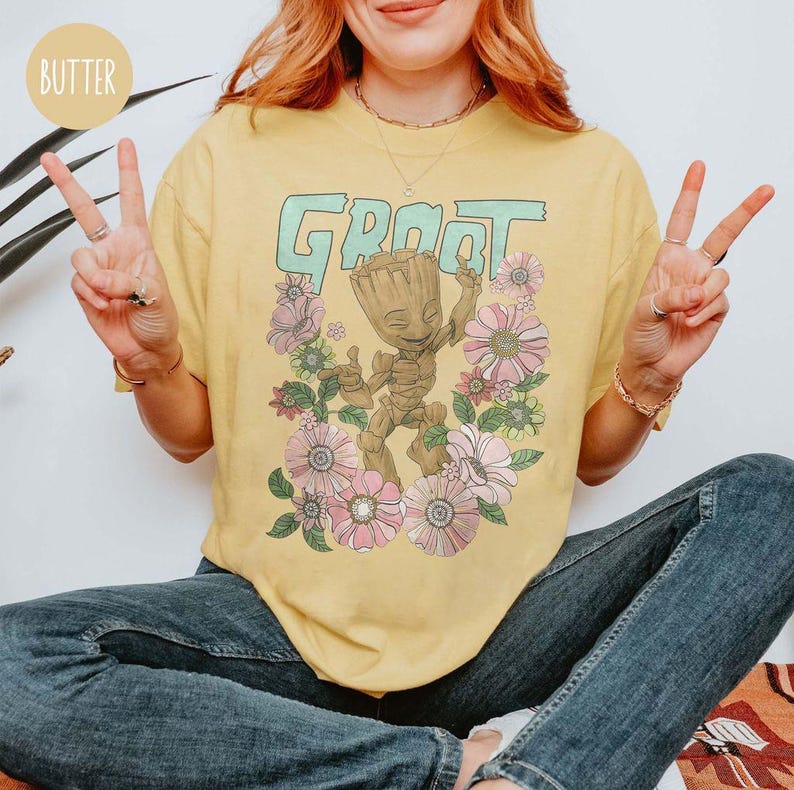 May include: A yellow t-shirt featuring a green "Groot" graphic and floral design. The graphic depicts a cartoon character with a green body and brown skin, standing in a field of flowers.