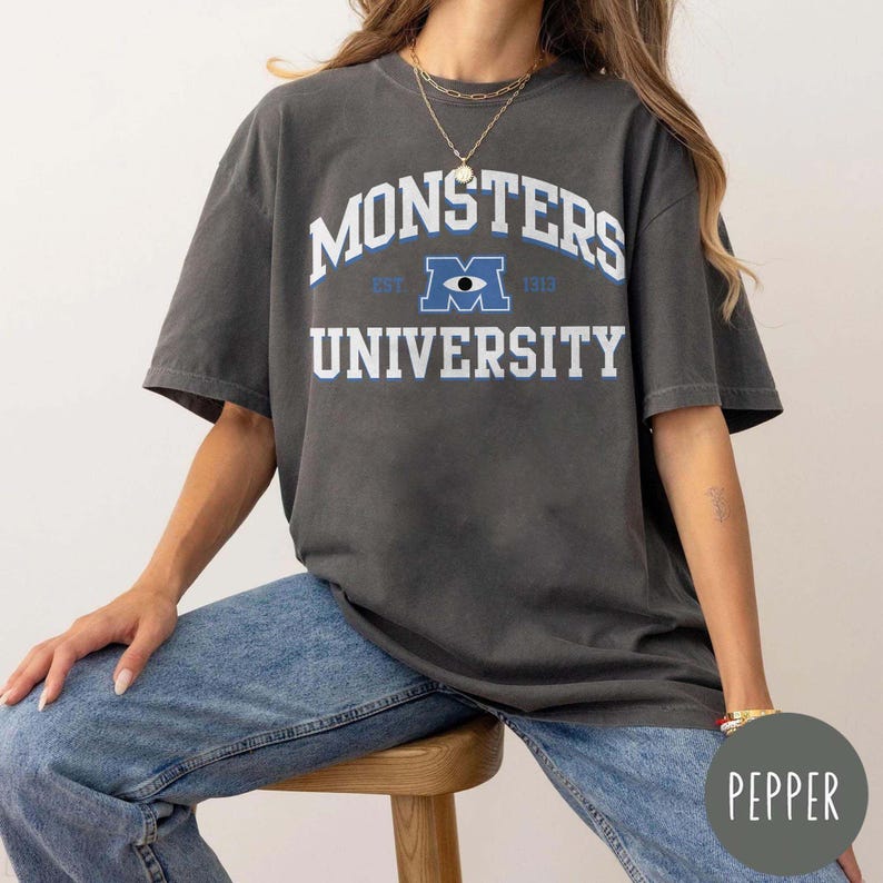 May include: Dark gray t-shirt with the text "MONSTERS UNIVERSITY" in white and blue. The shirt features a collegiate-style design with the letter "M" and an eye graphic. The shirt is made of a soft, comfortable material.