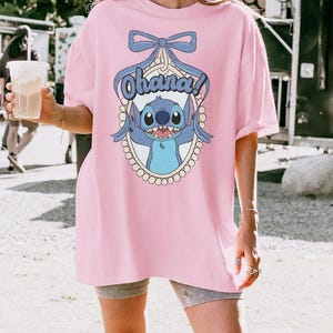Puede incluir: Una camiseta rosa de gran tama&ntilde;o con un gr&aacute;fico azul del personaje Stitch y el texto "Ohana!" en un marco ovalado blanco. El gr&aacute;fico incluye un lazo azul y un borde de perlas.