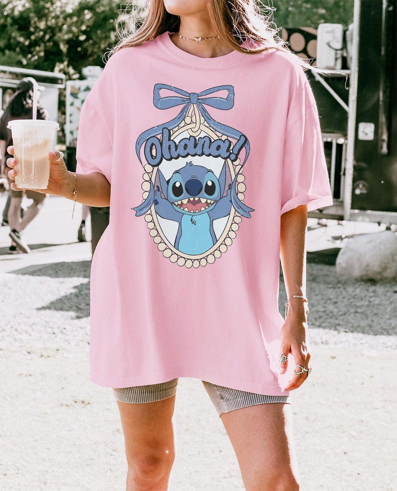 Puede incluir: Una camiseta rosa de gran tama&ntilde;o con un gr&aacute;fico azul del personaje Stitch y el texto "Ohana!" en un marco ovalado blanco. El gr&aacute;fico incluye un lazo azul y un borde de perlas.