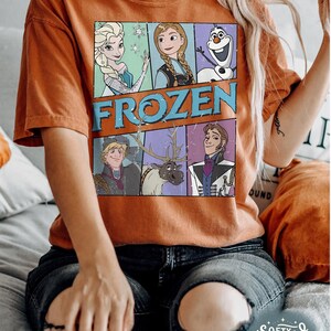 May include: A brown t-shirt with a vintage-style graphic of characters from the movie Frozen. The graphic includes Elsa, Anna, Olaf, Kristoff, Sven, and Hans. The text "FROZEN" is printed in a distressed font.