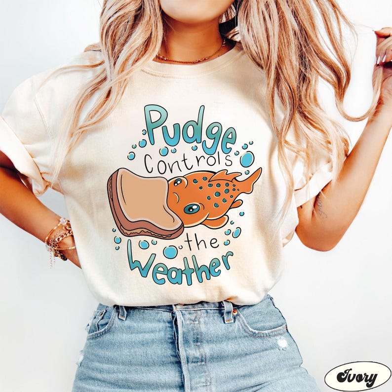 May include: A white t-shirt featuring a cartoon illustration of a fish wearing a peanut butter and jelly sandwich. The text on the shirt reads "Pudge Controls the Weather".