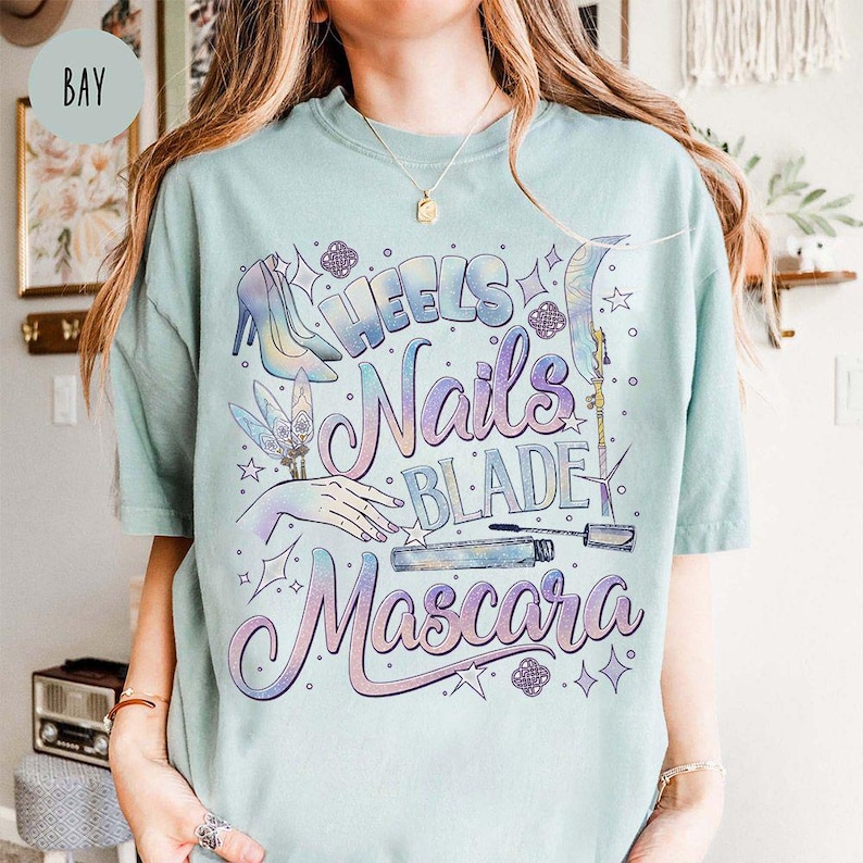 May include: A light blue t-shirt with a graphic design featuring the words "Heels, Nails, Blade, Mascara" in a cursive font. The design includes illustrations of high heels, a hand with manicured nails, a feather, and mascara.